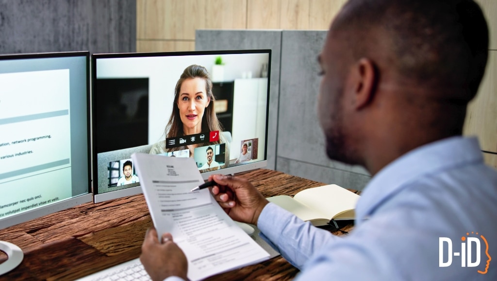 A man participates in a video conference on a dual-monitor setup, holding documents while speaking to colleagues and an interactive avatar visible on the screen.