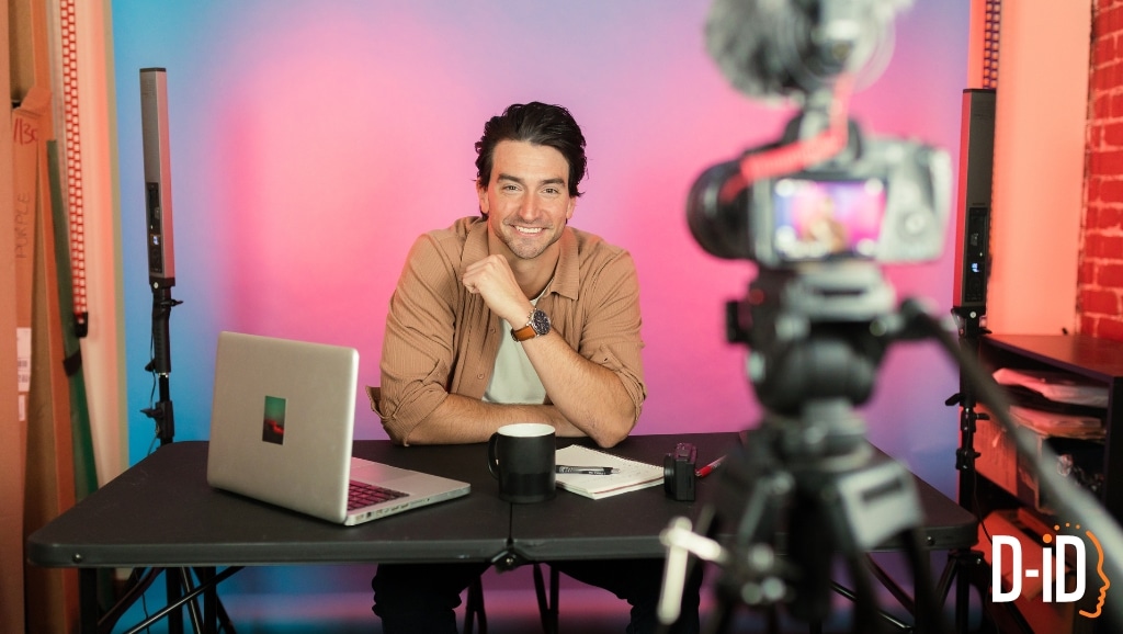 A man sits at a desk with a laptop and notebook, smiling at a video camera; two lights and the D-ID logo hint that he may be an ai generated influencer, set against a vibrant pink and blue background.