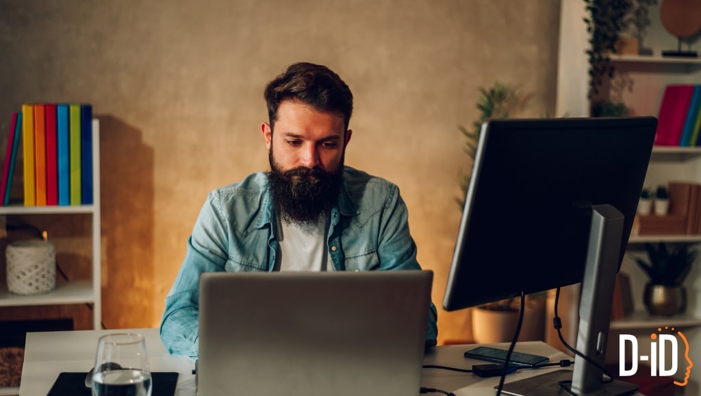 A man with a beard works at a desk with a laptop and monitor, surrounded by bookshelves filled with colorful books. The D-ID logo is visible in the bottom right, suggesting he's using an ai persona generator to create AI personas.