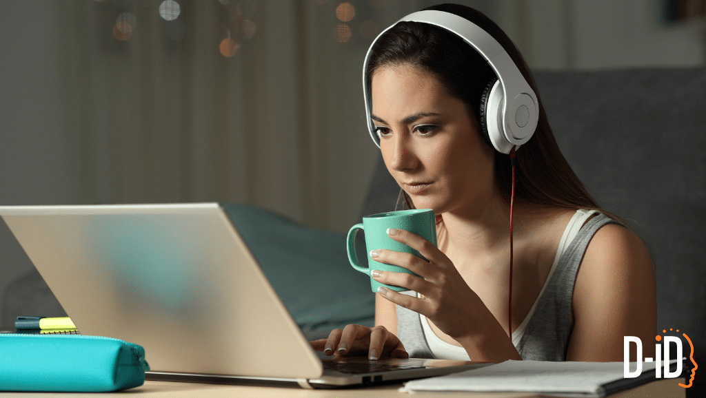 A woman wearing headphones sits at a desk using a laptop and holding a mug. A pencil case and notebook are nearby. The logo "D-ID" is visible in the corner, hinting she might be using a video tutorial maker to create a video tutorial.