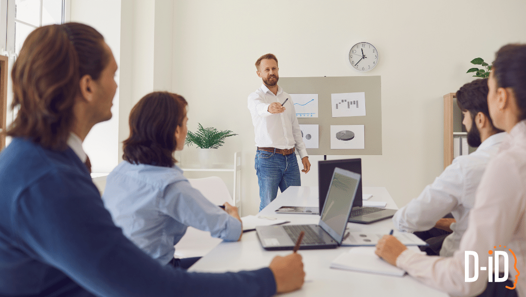 A man stands and presents data on charts to four seated colleagues in a meeting room; laptops are open on the table and a clock is on the wall, illustrating an effective sales training session.